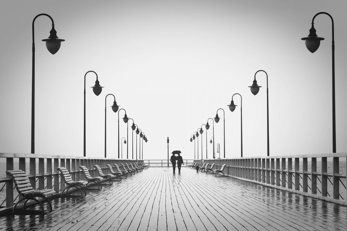 2 people walking down a pier at the seaside. Black and white image.