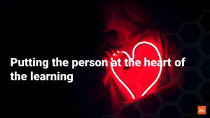 A person in darkness is hugging a red neon sign in the shape of a heart. The text says "putting teh person at the heart of learning"