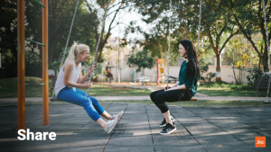 Two women sat on swings in a playpark talking to each other, face to face. The word on teh slide says "share"