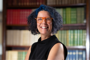 Head and shoulders shot of Dr Miriam firth, smiling broadly with a background of books on shelves. Her glasses are a fashionable red colour and her hair has blue highlights.