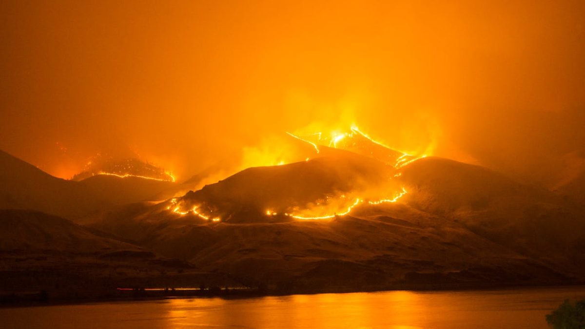 Nightime, a hillside by a lake is burning with extensive wildfirs. The fire is reflected in teh water and the air is full of smoke