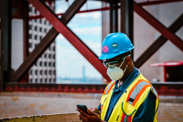 Construction worker in safety gear checking smartphone on a construction site.