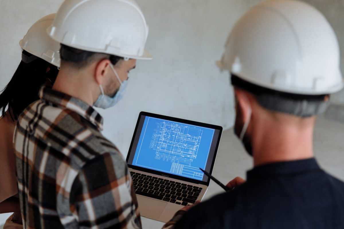 Engineers wearing hard hats and masks review a construction blueprint on a laptop indoors