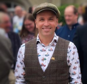A head and shoulders shot of Scott Hibberson. He is wearing a flat cap and is smiling broadly at the camera. There are people out of focus in the background.