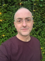 A head and shoulders shot of Chris Thomson. He wears glasses and has a on a burgundy t-shirt. He has short hair.