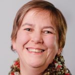 A head and shoulders shot of Lis Parcell. She is smiling broadly at the camera. She is wearing a colourful scarf and has short hair.